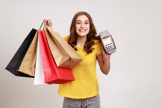 Positive smiling teenager girl with brown hair wearing yellow T-shirt holding payment terminal and paper shopping bags, easy express order and delivery. Indoor studio shot isolated on gray background.