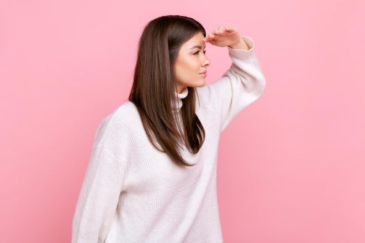 Side view portrait of attentive young woman looking far into distance, keeping hand over forehead, wearing white casual style sweater. Indoor studio shot isolated on pink background.