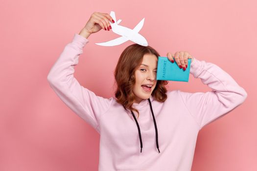 Portrait of happy curly haired teenage girl in hoodie winking, holding passport and paper airplane, looking at camera with toothy smile, travelling. Indoor studio shot isolated on pink background
