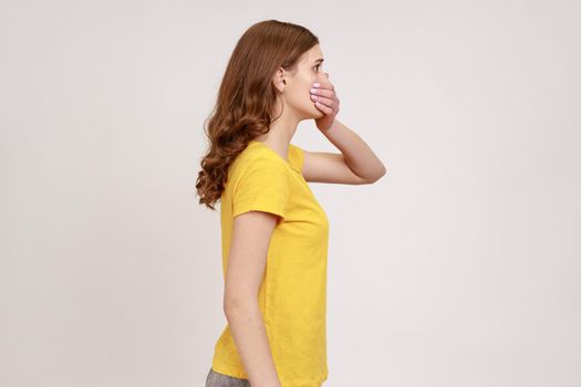 I will not say. Side view of scared bullied teen girl with brown hair in yellow t-shirt covering mouth with hand, keeping secret taboo. Indoor studio shot isolated on gray background.