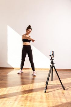 Coach conducting online sports training, pointing at phone camera on tripod, inviting you to workout, wearing black sports top and tights. Full length studio shot illuminated by sunlight from window.