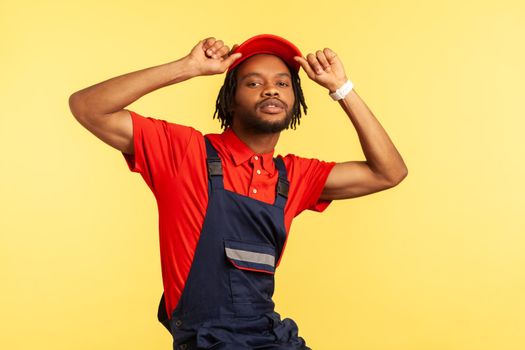 Portrait of stylish worker wearing blue uniform, red T-shirt posing with raised arms, keeps hands on his visor cap, looking directly at camera. Indoor studio shot isolated on yellow background.