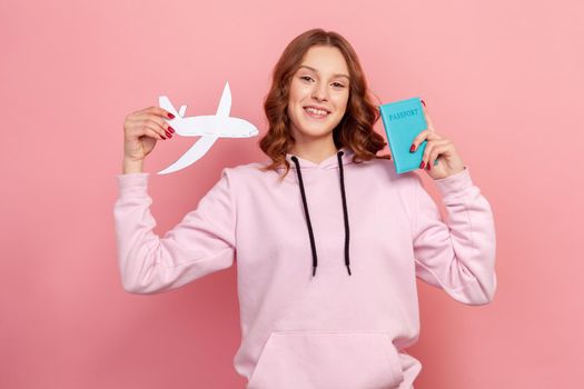 Portrait of joyful curly haired teenage girl in hoodie smiling and showing passport and paper airplane at camera, tourism. Indoor studio shot isolated on pink background