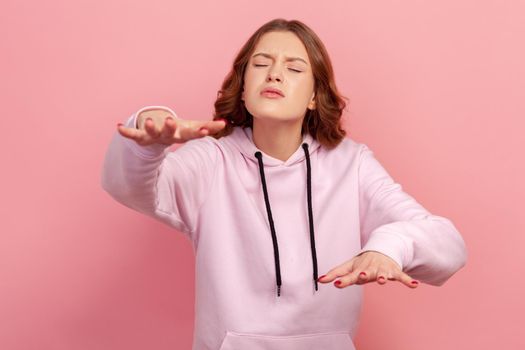 Portrait of concentrated brunette young female in hoodie trying to move with closed eyes and raised hands, vision problems, blindness. Indoor studio shot isolated on pink background