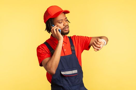 Portrait of concentrated courier wearing blue overalls talking phone with client, looking at smart watch, delivery service on time. Indoor studio shot isolated on yellow background.