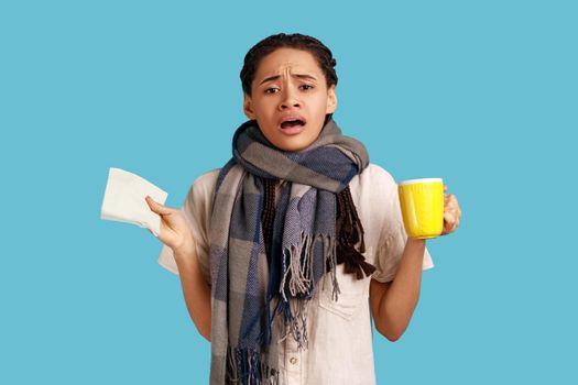 Unhealthy sick woman with black dreadlocks, wearing white shirt and wrapped in scarf, holding cup of tea, has influenza and runny nose. Indoor studio shot isolated on blue background.
