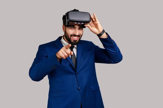 Smiling man with virtual reality glasses expressing happiness, being ready for playing game simulator, pointing to camera, choosing you as opponent. Indoor studio shot isolated on gray background.