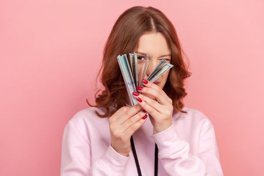 Portrait of curious curly haired teenage girl in hoodie peeping through bunch of dollar banknotes, passive income. Indoor studio shot isolated on pink background