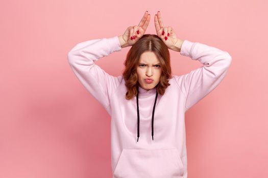 Portrait of aggressive teen girl in hoodie standing with bull horns, antler head gesture, threatening to attack, conflict or defence concept. Indoor studio shot isolated on pink background