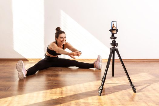 Female online trainer using phone on tripod for recording tutorials of stretching after workout, wearing black sports top and tights. Full length studio shot illuminated by sunlight from window.