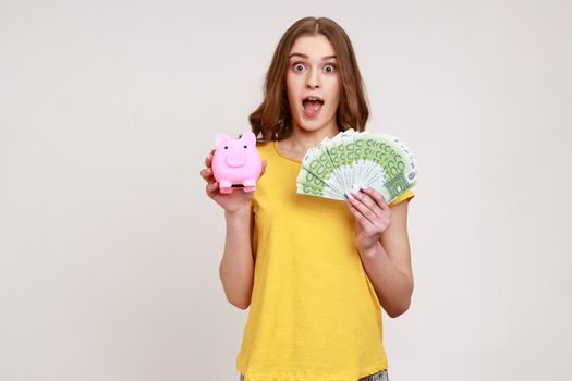 Extremely shocked female of young age in yellow T-shirt holding piggy bank and euro banknotes looking at camera with open mouth, deposit, cashback. Indoor studio shot isolated on gray background.