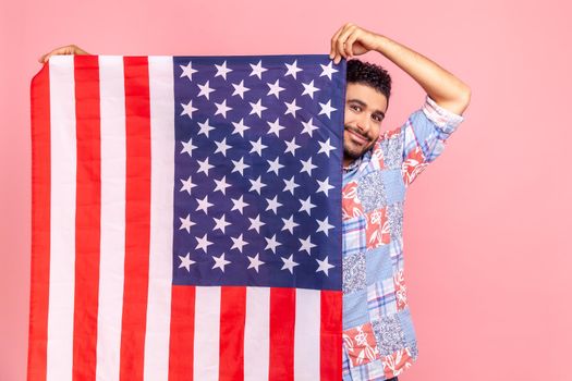 Portrait of attractive man holding USA flag in hands, looking at camera with toothy smile, proud of his social security in country, feeling in safe. Indoor studio shot isolated on pink background.