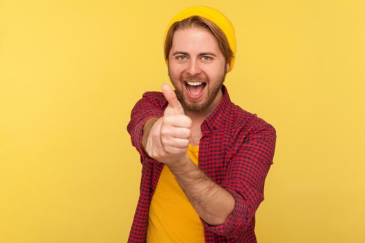 Portrait of hipster bearded guy in beanie hat standing with thumbs up, like gesture, demonstrating approval and agree with suggestion. Indoor studio shot isolated on yellow background.