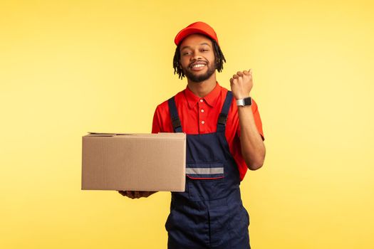 Pleasant looking deliveryman in overalls holds cardboard parcel and showing wristwatch, delivering in time, shipment and cargo transportation service. Indoor studio shot isolated on yellow background.