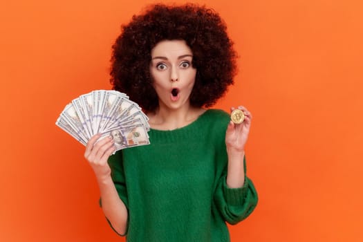 Attractive shocked woman with Afro hairstyle wearing green casual style sweater holding bitcoin and big sum of money, keeps mouth open, ecommerce. Indoor studio shot isolated on orange background.