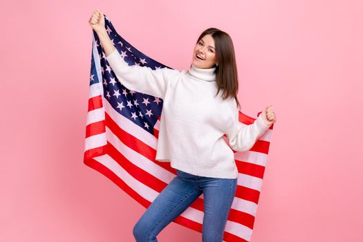 Portrait of pretty woman raised arms, holding american flag, celebrating national holiday, dancing, wearing white casual style sweater. Indoor studio shot isolated on pink background.