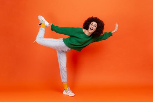 Full length portrait of funny woman with Afro hairstyle in green casual style sweater standing on one leg, pretending flying, yelling with excitement. Indoor studio shot isolated on orange background.