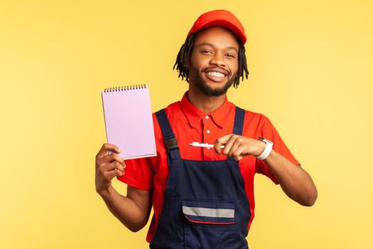 Portrait of workman wearing blue overalls and red cap, holding pointing at paper notebook with pleasant smile, presenting copy space. Indoor studio shot isolated on yellow background.