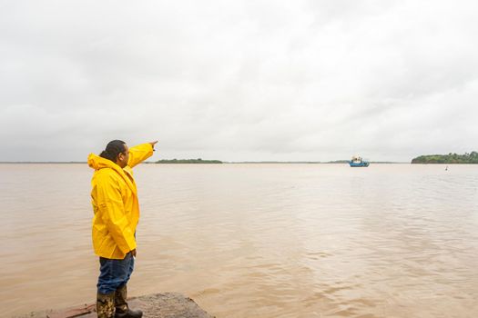 Latin hair mature man pointing with his hand to a boat in the sea and wearing raincoat on a dock in Bluefields Nicaragua