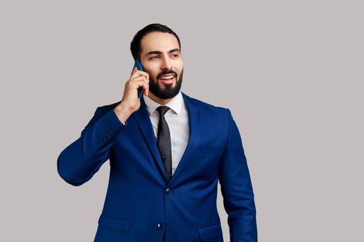 Smiling man standing, talking smartphone, making call and having pleasant conversation with friend or business partner, wearing official style suit. Indoor studio shot isolated on gray background.