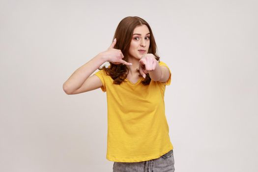 Portrait of beautiful brown haired teenager girl in casual yellow T-shirt standing with call gesture, looking and pointing at camera. Indoor studio shot isolated on gray background.