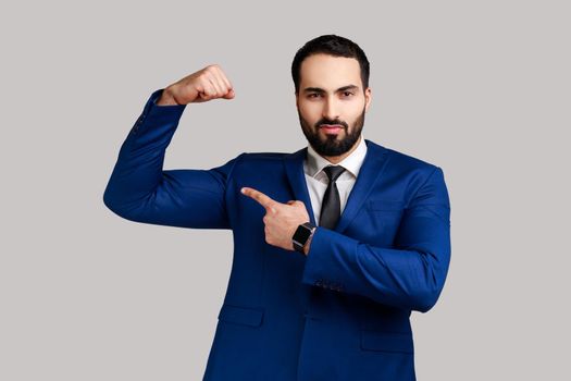 Strong bearded man raising hands showing power, pointing at his biceps, feeling independent strong with proud look, wearing official style suit. Indoor studio shot isolated on gray background.