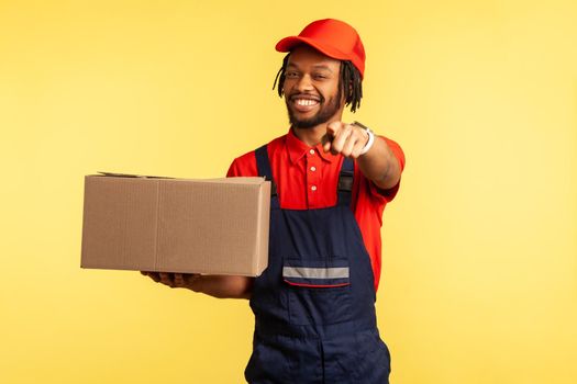 Professional delivery man in uniform holding cardboard boxes and pointing to camera, choosing you, shipment and cargo transportation service. Indoor studio shot isolated on yellow background.