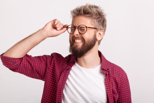 Portrait of funny extremely happy bearded man touching eyeglasses standing with closed eyes and laughing. Indoor studio shot isolated on gray background