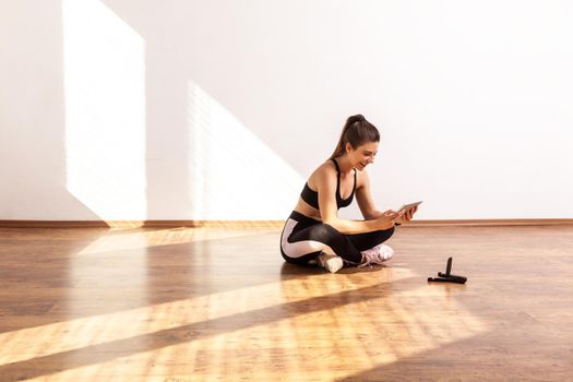 Girl sitting on floor with tablet, searching tutorial video for sport exercising, workout at home, wearing black sports top and tights. Full length studio shot illuminated by sunlight from window.