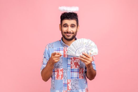Hey you, make money. Portrait of positive angelic bearded man with halo over head in blue shirt holding fan of dollar bills, pointing finger to camera. Indoor studio shot isolated on pink background.