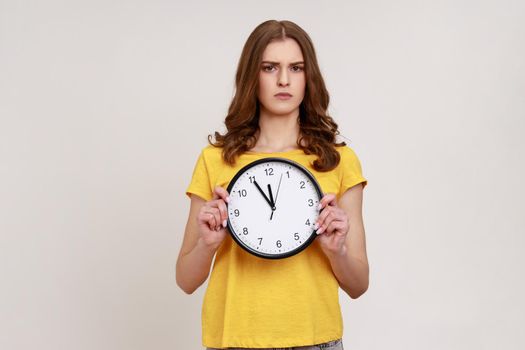 Serious strict teenager girl with wavy hair in casual yellow T-shirt afraid of being late, holding in hand wall watch, deadline, punctuality. Indoor studio shot isolated on gray background.