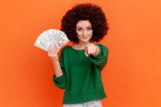 Hey you. Rich satisfied woman with Afro hairstyle wearing green casual style sweater holding fan of dollars banknotes and pointing finger to camera. Indoor studio shot isolated on orange background.