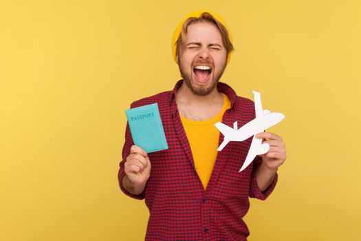 Portrait of crazy excited hipster bearded guy in beanie hat and checkered shirt holding passport document and airplane mockup, rejoicing travel tour. Indoor studio shot isolated on yellow background.