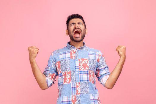 Portrait of overjoyed handsome man with beard in casual shirt expressing winning gesture with raised fists and screaming, celebrating victory. Indoor studio shot isolated on pink background.