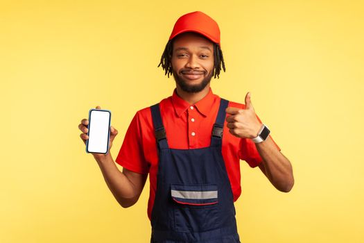 Satisfied handyman wearing overalls holding smart phone with blank screen for advertisement, showing thumb up, recommend service industry online app. Indoor studio shot isolated on yellow background.