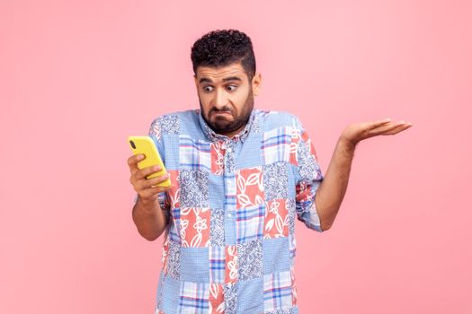 Portrait of attractive bearded man wearing blue casual shirt holding smart phone in hands, spreading hans aside, dont know how to use application. Indoor studio shot isolated on pink background.