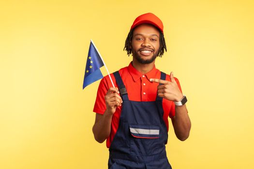 Happy handyman in uniform with dreadlocks pointing finger at Europe union flag in his hand and looking at camera with positive expression. Indoor studio shot isolated on yellow background.