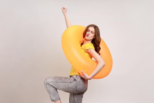 Portrait of carefree playful attractive woman of young age in yellow T-shirt and jeans raising hand, poses with rubber ring, smiling at camera. Indoor studio shot isolated on gray background.