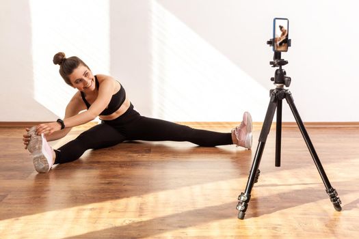 Girl coach stretching legs, broadcasting online class, using phone on tripod for recording video, wearing black sports top and tights. Full length studio shot illuminated by sunlight from window.