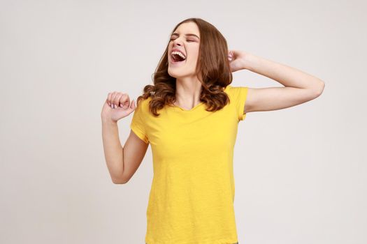 Portrait of beautiful young woman with brown wavy hair in yellow casual T-shirt standing and yawning with closed eyes and raised arms. Indoor studio shot isolated on gray background.