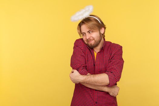 Portrait of hipster bearded guy in checkered shirt and with nimbus overhead hugging himself with love, worship to beauty, over-inflated ego. Indoor studio shot isolated on yellow background.