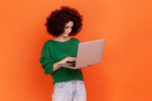 Portrait of attractive concentrated woman with Afro hairstyle wearing green casual style sweater holding laptop, freelancer working online. Indoor studio shot isolated on orange background.