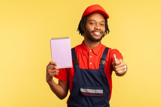 Portrait of optimistic mechanic or handyman wearing blue overalls and red T-shirt, showing paper notebook and holding out pen, copy space. Indoor studio shot isolated on yellow background.