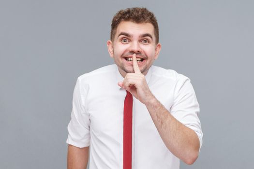 this is secret. Portrait of young handsome man in white shirt and tie standing with finger on lips, tell secret and smiling. indoor isolated on gray background.