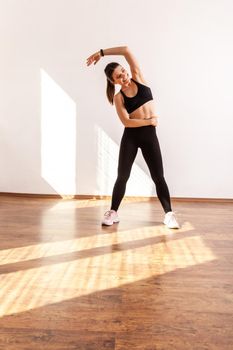 Slim female raising hand bending to side, doing stretching muscle workouts, flexibility exercises, wearing black sports top and tights. Full length studio shot illuminated by sunlight from window.
