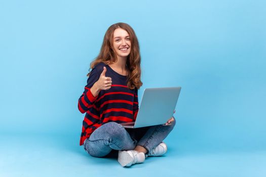 Smiling woman wearing striped casual style sweater, sitting with laptop on floor, working online, showing thumb up, satisfied with teleworking. Indoor studio shot isolated on blue background.