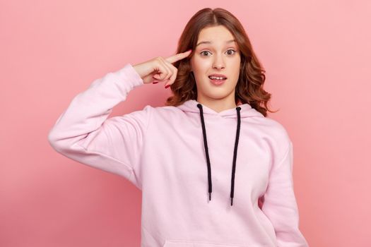Portrait of teen girl in hoodie showing stupid gesture with finger near head, out of mind, accusing crazy dumb plan, reckless expression. Indoor studio shot isolated on pink background