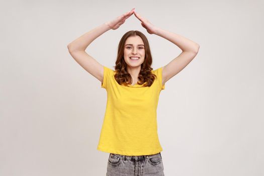I'm in safety. Portrait of happy teenager girl with wavy hair in yellow t-shirt standing and raising hands, showing roof gesture, dreaming of house. Indoor studio shot isolated on gray background.