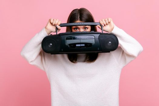 Portrait of shy brunette female holds tape recorder, hiding her face with record player, peeping out, wearing white casual style sweater. Indoor studio shot isolated on pink background.