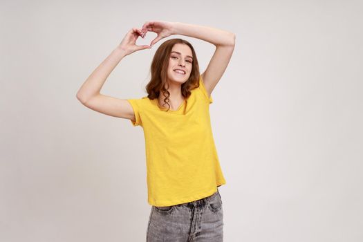 Happy beautiful teenager girl with brown wavy hair wearing T-shirt raising hands and showing heart shape gesture, flirting, demonstrating affection. Indoor studio shot isolated on gray background.